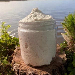 May include: A clear glass jar filled with a thick, white, whipped body scrub. The jar is sitting on a tree stump with a body of water in the background.