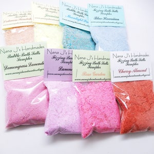 May include: A variety pack of bath salts in clear plastic bags with labels. The labels are white with black text and list the scents: Lemongrass Lavender, Lavender, Rose Garden, and Cherry Almond. The bath salts are pink, light pink, and orange.