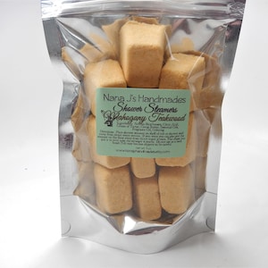 May include: A clear, sealed bag filled with rectangular, light brown shower steamers. A green label reads "Nana J's Handmades Shower Steamers Mahogany Teakwood." The net weight is 3 oz. The bag is on a white background.
