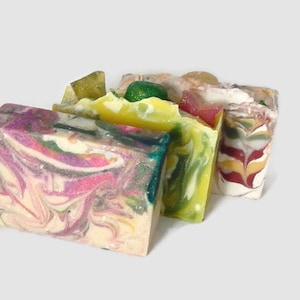 May include: Three bars of handmade soap with colorful swirls and patterns. The soap bars are stacked on top of each other. The top bar is a yellow and green swirl with a green leaf on top. The middle bar is a green and white swirl. The bottom bar is a pink, white, and green swirl.