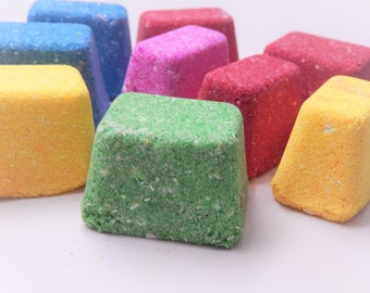May include: A collection of multi-colored, trapezoidal bath bombs in vibrant shades of green, yellow, blue, pink, and red. Each solid block features a textured, granular surface with visible white flecks throughout the compressed material.