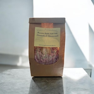 May include: A brown paper bag with a clear window, filled with lavender and chamomile bath salts. The label reads "Nana J's Handmade Premium Bubble Bath Salts Lavender & Chamomile". The bag sits on a light-colored surface.