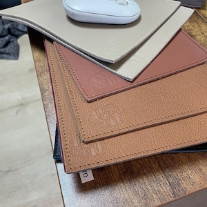 May include: A stack of five leather mouse mats in various shades of brown, beige, and red. The top mouse mat has a white wireless mouse on it. The mouse mats have a stitched edge and a subtle embossed design.