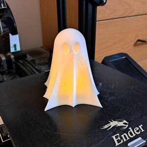 Puede incluir: Una figura de fantasma blanca impresa en 3D con dos agujeros para los ojos. El fantasma está iluminado desde adentro, proyectando un brillo cálido. El fantasma está sentado sobre una superficie negra con la palabra "Ender" impresa en ella.