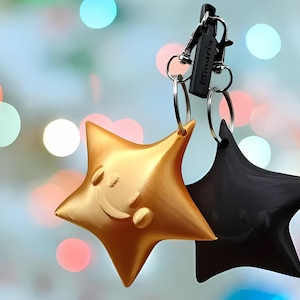 Puede incluir: Un llavero en forma de estrella dorada y negra con una cara sonriente en la estrella dorada. El llavero está unido a un anillo plateado y a un clip de metal negro con la palabra "Tamaris" impresa en él.