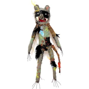 May include: A handmade raccoon doll with a distressed aesthetic. The doll has a black mask, red mouth, and stitched details. Its body is a patchwork of green, grey, and brown fabrics, with black fur accents and threadbare limbs.