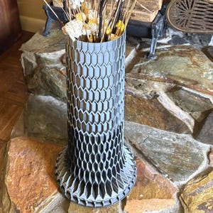 Puede incluir: Un jarrón alto y gris con un patrón de panal de abeja contiene un arreglo decorativo. El arreglo incluye ramas negras y bronceadas y adornos blancos en forma de flor. El jarrón está sobre una superficie de piedra.