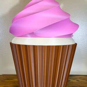 K&ouml;nnte beinhalten: Eine gro&szlig;e, dekorative Cupcake-Skulptur mit einer rosa, gewirbelten Glasur und einem braunen, geriffelten Boden. Die Glasur hat einen Farbverlauf von Rosat&ouml;nen, und der Boden ist dunkelbraun. Der Cupcake steht auf einer Holzoberfl&auml;che.