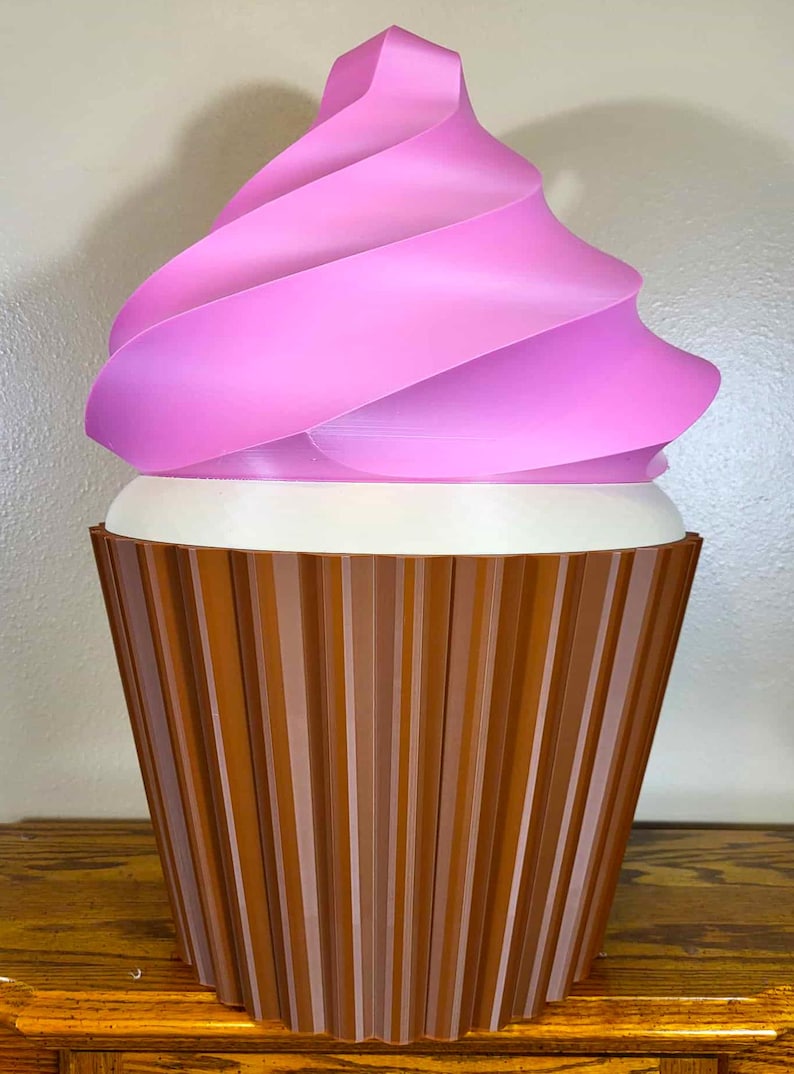 K&ouml;nnte beinhalten: Eine gro&szlig;e, dekorative Cupcake-Skulptur mit einer rosa, gewirbelten Glasur und einem braunen, geriffelten Boden. Die Glasur hat einen Farbverlauf von Rosat&ouml;nen, und der Boden ist dunkelbraun. Der Cupcake steht auf einer Holzoberfl&auml;che.