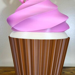 K&ouml;nnte beinhalten: Eine gro&szlig;e, dekorative Cupcake-Skulptur. Das rosa Frosting wirbelt &uuml;ber einer wei&szlig;en Basis, mit einer braunen, geriffelten Cupcake-Form. Die Skulptur wird auf einer Holzoberfl&auml;che ausgestellt.