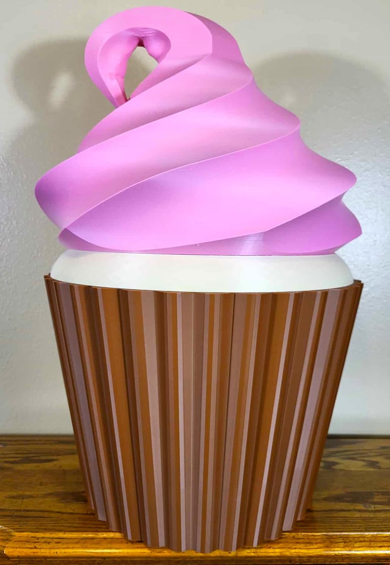 K&ouml;nnte beinhalten: Eine gro&szlig;e, dekorative Cupcake-Skulptur. Das rosa Frosting wirbelt &uuml;ber einer wei&szlig;en Basis, mit einer braunen, geriffelten Cupcake-Form. Die Skulptur wird auf einer Holzoberfl&auml;che ausgestellt.