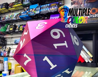 Può includere: Un grande dado a venti facce multicolore con numeri bianchi. Il dado è principalmente viola e rosa, con il testo "MULTIPLE COLOR CHOICES" sullo sfondo. L'immagine è scattata in un negozio, con scaffali di giochi e carte collezionabili visibili.
