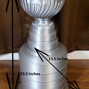 Może przedstawiać: Srebrne trofeum z żłobionym wierzchem i warstwową podstawą. Trofeum ma 64,8 cm wysokości i 34,3 cm szerokości. Projekt zawiera ułożone w stos pierścienie i dekoracyjne detale.