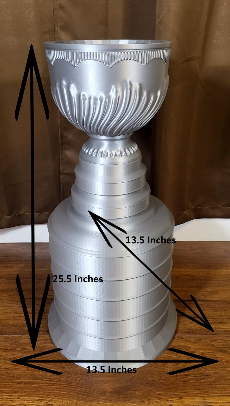 Może przedstawiać: Srebrne trofeum z żłobionym wierzchem i warstwową podstawą. Trofeum ma 64,8 cm wysokości i 34,3 cm szerokości. Projekt zawiera ułożone w stos pierścienie i dekoracyjne detale.