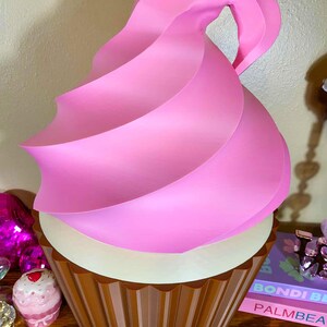 K&ouml;nnte beinhalten: Eine gro&szlig;e, dekorative Cupcake-Skulptur mit einem braunen, geriffelten Sockel und einem wei&szlig;en Mittelteil. Die Oberseite zeigt einen Wirbel aus rosa Zuckerguss. Die Skulptur ist als riesiger Cupcake gestaltet.