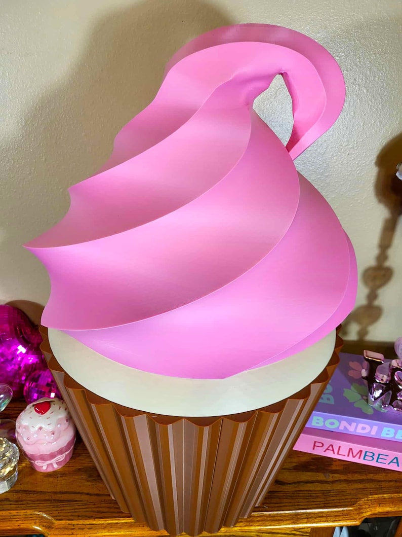 K&ouml;nnte beinhalten: Eine gro&szlig;e, dekorative Cupcake-Skulptur mit einem braunen, geriffelten Sockel und einem wei&szlig;en Mittelteil. Die Oberseite zeigt einen Wirbel aus rosa Zuckerguss. Die Skulptur ist als riesiger Cupcake gestaltet.