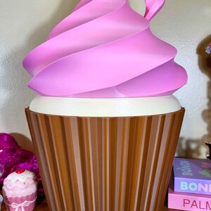 K&ouml;nnte beinhalten: Ein gro&szlig;er, dekorativer Aufbewahrungsbeh&auml;lter in Cupcake-Form. Der Beh&auml;lter hat einen braunen, gerippten Boden, einen wei&szlig;en Mittelteil und einen rosa Wirbel oben. Das Design &auml;hnelt einem riesigen Cupcake.