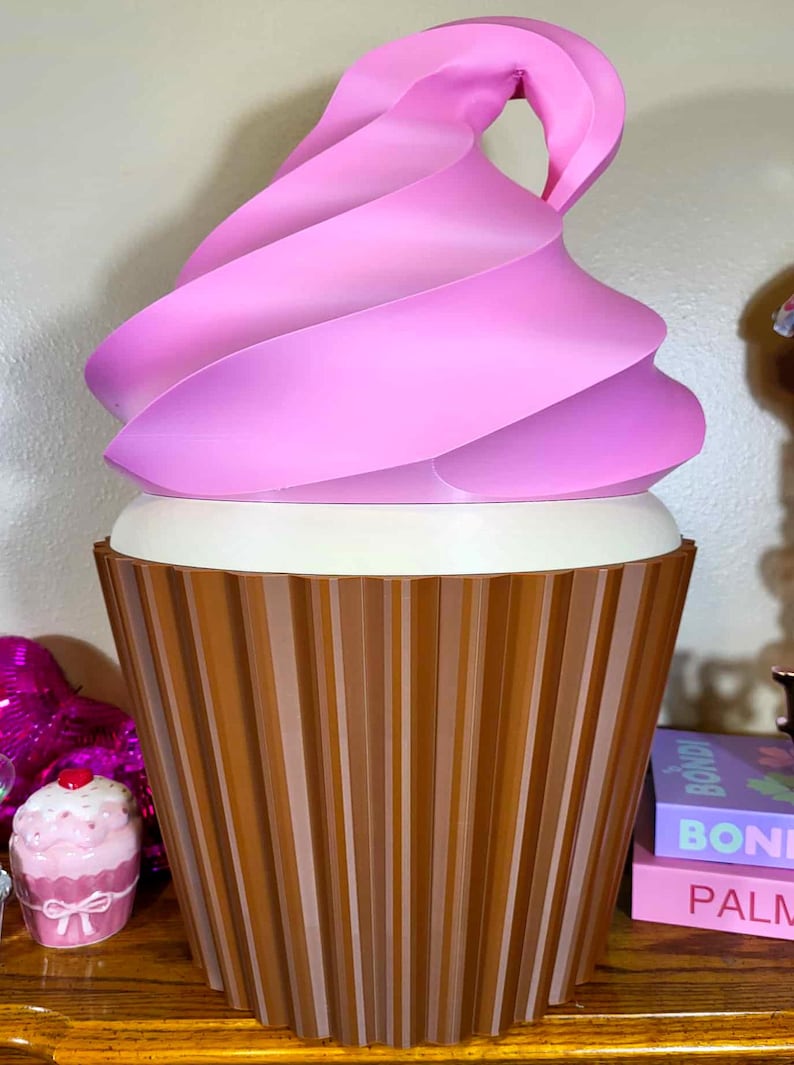 K&ouml;nnte beinhalten: Ein gro&szlig;er, dekorativer Aufbewahrungsbeh&auml;lter in Cupcake-Form. Der Beh&auml;lter hat einen braunen, gerippten Boden, einen wei&szlig;en Mittelteil und einen rosa Wirbel oben. Das Design &auml;hnelt einem riesigen Cupcake.