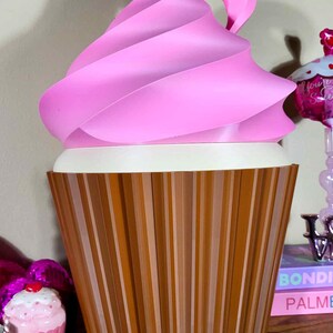 K&ouml;nnte beinhalten: Eine gro&szlig;e, dekorative Cupcake-Skulptur mit einem rosa, gewirbelten Oberteil und einer braunen, vertikal gestreiften Basis. Der wei&szlig;e Mittelteil kontrastiert mit Rosa und Braun. Die Skulptur ist eine skurrile, &uuml;bergro&szlig;e Darstellung eines Cupcakes.