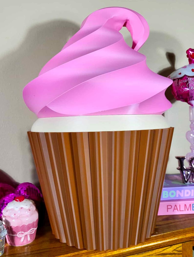 K&ouml;nnte beinhalten: Eine gro&szlig;e, dekorative Cupcake-Skulptur mit einem rosa, gewirbelten Oberteil und einer braunen, vertikal gestreiften Basis. Der wei&szlig;e Mittelteil kontrastiert mit Rosa und Braun. Die Skulptur ist eine skurrile, &uuml;bergro&szlig;e Darstellung eines Cupcakes.