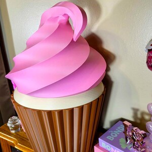 K&ouml;nnte beinhalten: Eine gro&szlig;e, dekorative Cupcake-Skulptur mit rosa Wirbelfrosting. Der Cupcake hat eine braune, gerippte Basis und eine wei&szlig;e Cremef&uuml;llung. Die Skulptur steht auf einer Holzoberfl&auml;che.