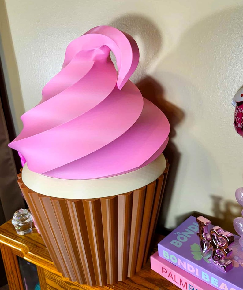 K&ouml;nnte beinhalten: Eine gro&szlig;e, dekorative Cupcake-Skulptur mit rosa Wirbelfrosting. Der Cupcake hat eine braune, gerippte Basis und eine wei&szlig;e Cremef&uuml;llung. Die Skulptur steht auf einer Holzoberfl&auml;che.