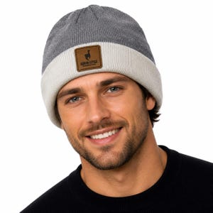 May include: A two-tone knit beanie featuring a dark gray crown and a light gray folded cuff. A square brown leather patch is centered on the cuff, displaying a black silhouette of an alpaca and the text ANDEAN ALPACA HANDMADE CLOTHING.