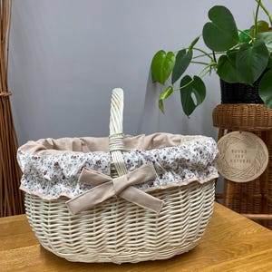 Può includere: Un cestino in vimini color crema con manico e fodera in tessuto floreale. Un fiocco beige è attaccato sul davanti. Il cestino è su una superficie di legno. Un cartello con la scritta "Round and Natural" è visibile.