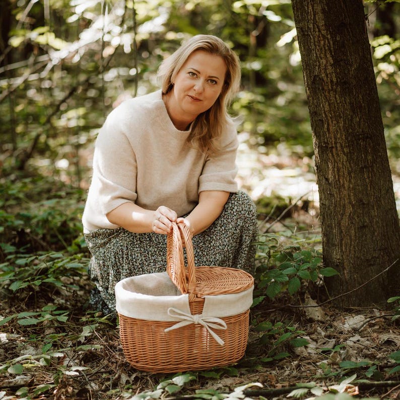 May include: A woman in a forest setting is holding a wicker picnic basket with a white linen lining. The basket is open and the lid is resting on the ground.