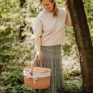May include: A woman in a floral skirt holds a wicker picnic basket with a white fabric lining and a brown ribbon.