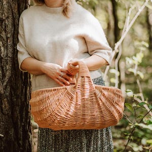 May include: A woman holds a large, woven wicker basket with a handle. The basket is light brown and has a rounded shape. The woman is wearing a light beige sweater and a floral skirt. The background is a blurred image of a forest.