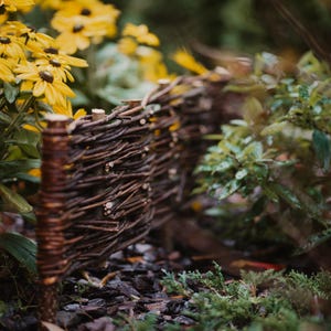 Könnte beinhalten: Ein geflochtener brauner Zweigzaun in einer Gartenlandschaft. Gelbe Blumen sind im Vordergrund.