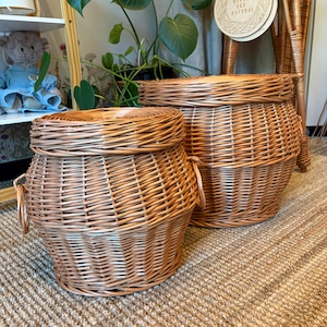 Op de afbeelding: Twee ronde, lichtbruine gevlochten rieten manden met bijpassende deksels en handvatten aan de zijkanten. De manden hebben een taps toelopende basis en een breder middengedeelte, gemaakt van natuurlijke wilgentenen.