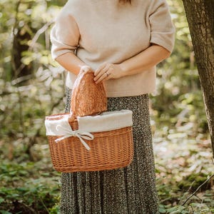 May include: A wicker picnic basket with a white fabric lining and a brown wicker lid. The basket is being held by a person wearing a beige sweater and a floral skirt.