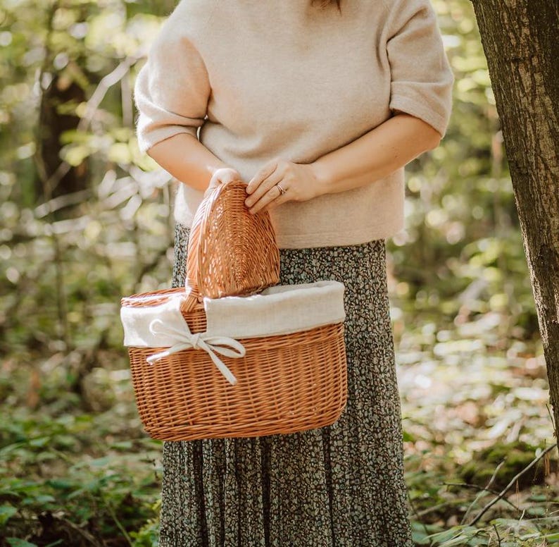 May include: A wicker picnic basket with a white fabric lining and a brown wicker lid. The basket is being held by a person wearing a beige sweater and a floral skirt.