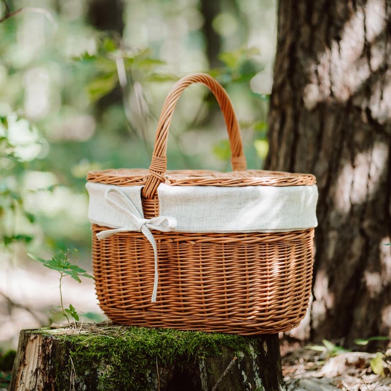 May include: A brown wicker picnic basket with a white fabric lid and a white ribbon. The basket is sitting on a tree stump in a forest.