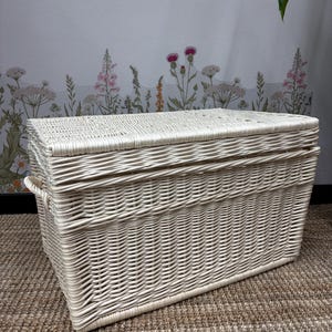 Pode incluir: Um grande baú de vime branco sujo com formato retangular e tampa articulada. O baú apresenta detalhes tecidos e alças laterais. O baú está colocado sobre um tapete tecido castanho, com um papel de parede floral ao fundo.