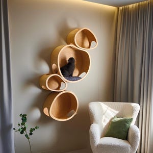 May include: A modern cat furniture set featuring five wooden, circular wall-mounted cat beds. A black cat is resting in one of the beds. A white armchair with a green pillow is in the foreground. The room has light-colored walls and flooring.