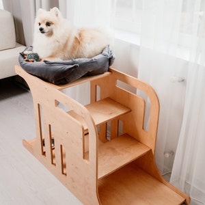 Puede incluir: Una rampa de madera de color marrón claro para mascotas con tres escalones y una plataforma acolchada en la parte superior. Un perro pequeño blanco está acostado en la plataforma.