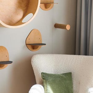 Peut inclure: Ensemble de meubles pour chats en bois, comprenant plusieurs étagères murales et un lit circulaire. Les étagères ont une finition en bois naturel avec une surface en feutre gris foncé. Un fauteuil blanc avec un coussin vert est au premier plan.