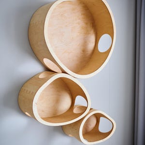 May include: Three wooden wall shelves in varying sizes, mounted on a light gray wall. The shelves are made of light-colored wood and have organic, rounded shapes with cut-out openings. The shelves are designed to be decorative and functional.