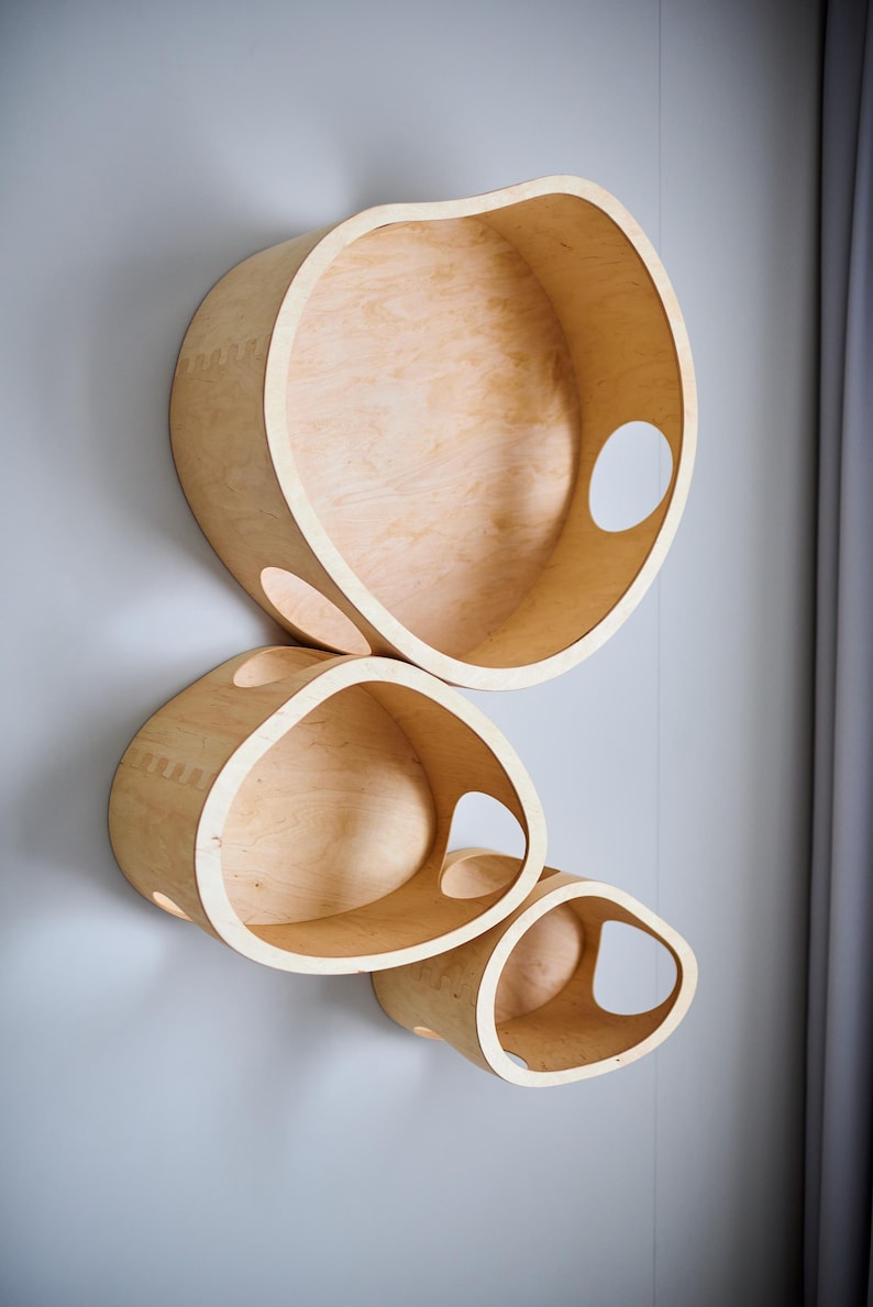 May include: Three wooden wall shelves in varying sizes, mounted on a light gray wall. The shelves are made of light-colored wood and have organic, rounded shapes with cut-out openings. The shelves are designed to be decorative and functional.