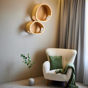 May include: Two wooden, oval-shaped wall shelves with a natural finish are mounted on a gray wall. A white armchair with a green pillow and blanket sits nearby. A small vase with greenery is on the floor.