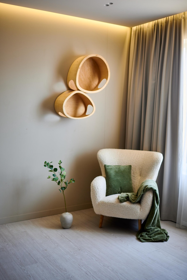 May include: Two wooden, oval-shaped wall shelves with a natural finish are mounted on a gray wall. A white armchair with a green pillow and blanket sits nearby. A small vase with greenery is on the floor.
