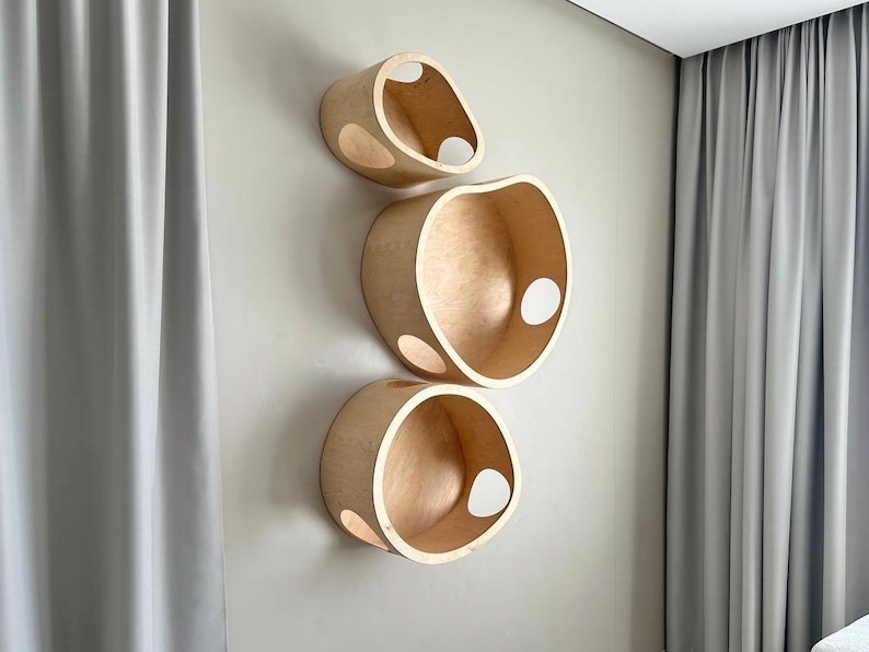 May include: Three wooden, wall-mounted shelves with a natural finish. Each shelf has a unique, rounded shape with a large opening and several smaller, circular cutouts. The shelves are arranged vertically on a light-colored wall.