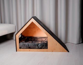 Op de afbeelding: Een houten hondenhuis met een zwart dak en een grijs kussen erin. Het huis is gemaakt van licht hout en heeft een driehoekige vorm.