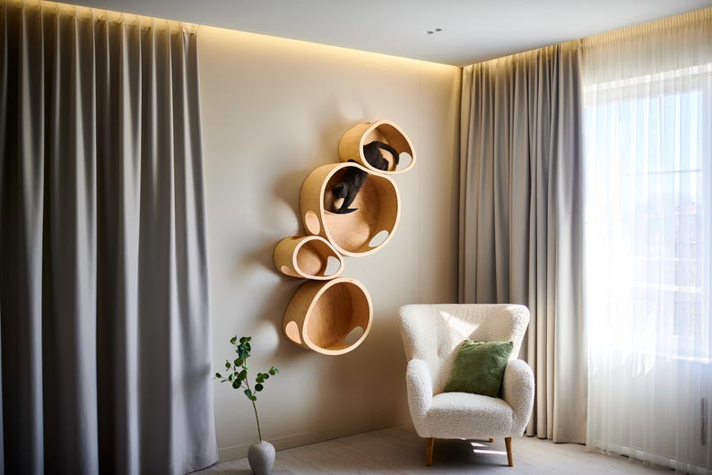 May include: A modern cat furniture set with multiple circular wooden shelves mounted on a beige wall. Two black cats are resting in the shelves. A white armchair with a green pillow sits nearby. The room has beige curtains and a plant in a vase.