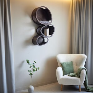 May include: A modern wall-mounted cat shelf unit with three oval-shaped compartments in dark brown. The top shelf has a gray cushion. A white armchair with a green pillow and blanket sits nearby. A vase with greenery is in the foreground.