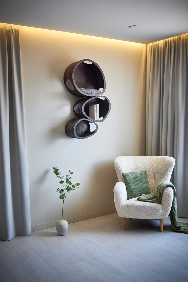 May include: A modern wall-mounted cat shelf unit with three oval-shaped compartments in dark brown. The top shelf has a gray cushion. A white armchair with a green pillow and blanket sits nearby. A vase with greenery is in the foreground.