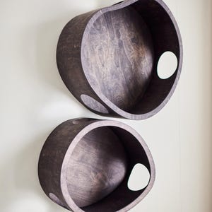 May include: Two dark brown wooden wall shelves with a unique, curved design. Each shelf has a circular opening on the side. The shelves are mounted on a light-colored wall, showcasing their modern aesthetic.
